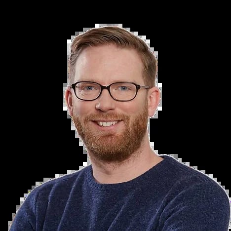 Smiling man with glasses and reddish-brown beard wearing a dark blue sweater, professional headshot on black background